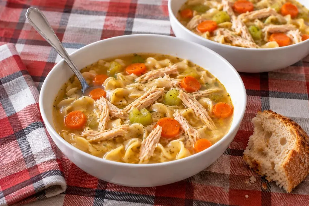 Two bowls of chicken noodle soup on a checkered tablecloth with a spoon and a piece of crusty bread
