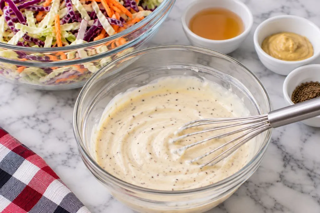 Creamy coleslaw dressing whisked in a glass bowl with visible flecks of celery seed and black pepper