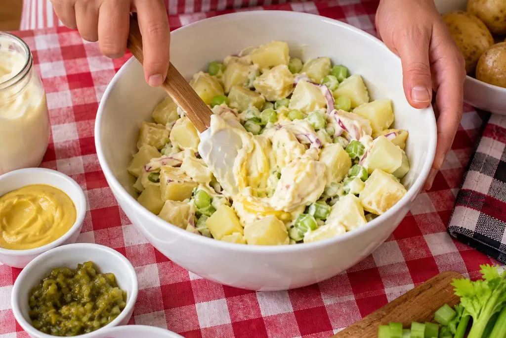 Hands folding mayonnaise dressing into warm cubed potatoes, peas, celery and red onion for classic potato salad