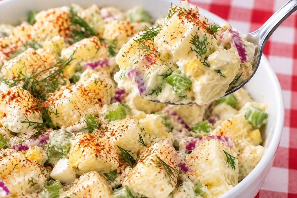 Spoon lifting a scoop of creamy classic potato salad with peas, celery and red onion from the bowl
